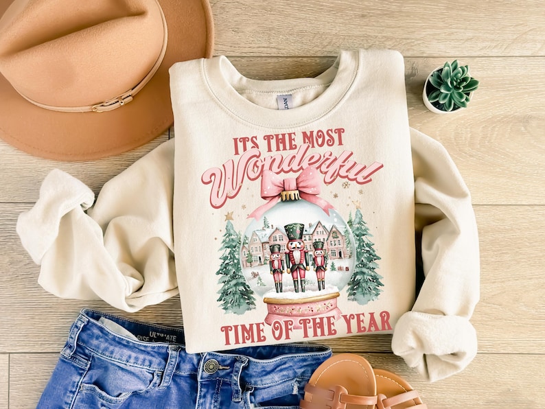 May include: A cream-coloured sweatshirt with a Christmas-themed design. The design features a snow globe with three nutcracker figures inside, surrounded by houses and trees. The text "It's the most Wonderful Time of the Year" is printed below the snow globe.