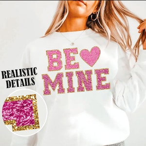 Puede incluir: Sudadera blanca con letras de purpurina rosa que dicen "BE MINE" con un corazón de purpurina rosa.