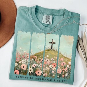 May include: A teal blue t-shirt with a graphic print of a cross on a hill with flowers. The text "NOTHING IS IMPOSSIBLE WITH GOD" is printed on the bottom of the shirt.