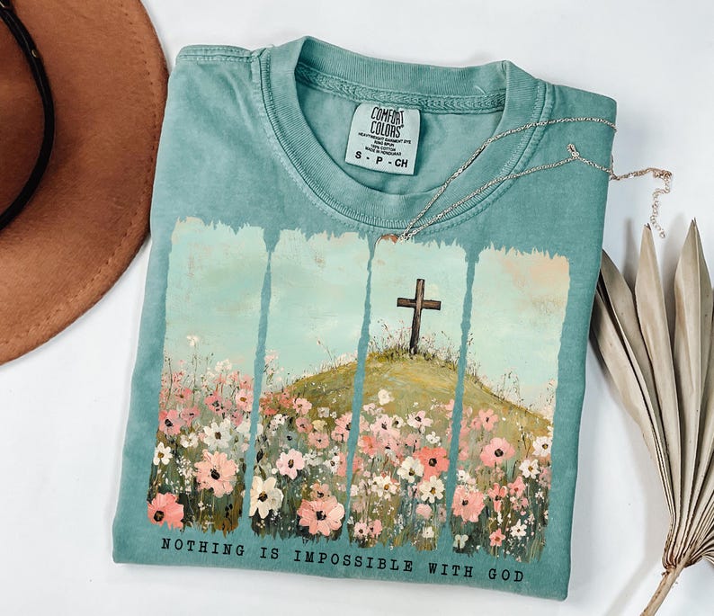 May include: A teal blue t-shirt with a graphic print of a cross on a hill with flowers. The text "NOTHING IS IMPOSSIBLE WITH GOD" is printed on the bottom of the shirt.