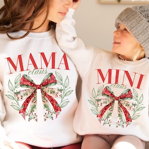 May include: White sweatshirts with matching designs. One sweatshirt reads "MAMA claus" in red, and the other reads "MINI claus" in red. Both feature a watercolor bow with red, green, and white accents, surrounded by green leaves. A festive holiday theme.