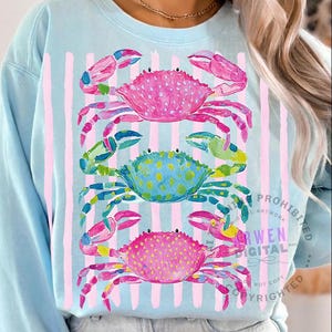 May include: Light blue sweatshirt featuring a watercolor design of three colorful crabs stacked vertically. The crabs are pink, blue, and pink again, with pink and green accents. Vertical pink stripes are behind the crabs.