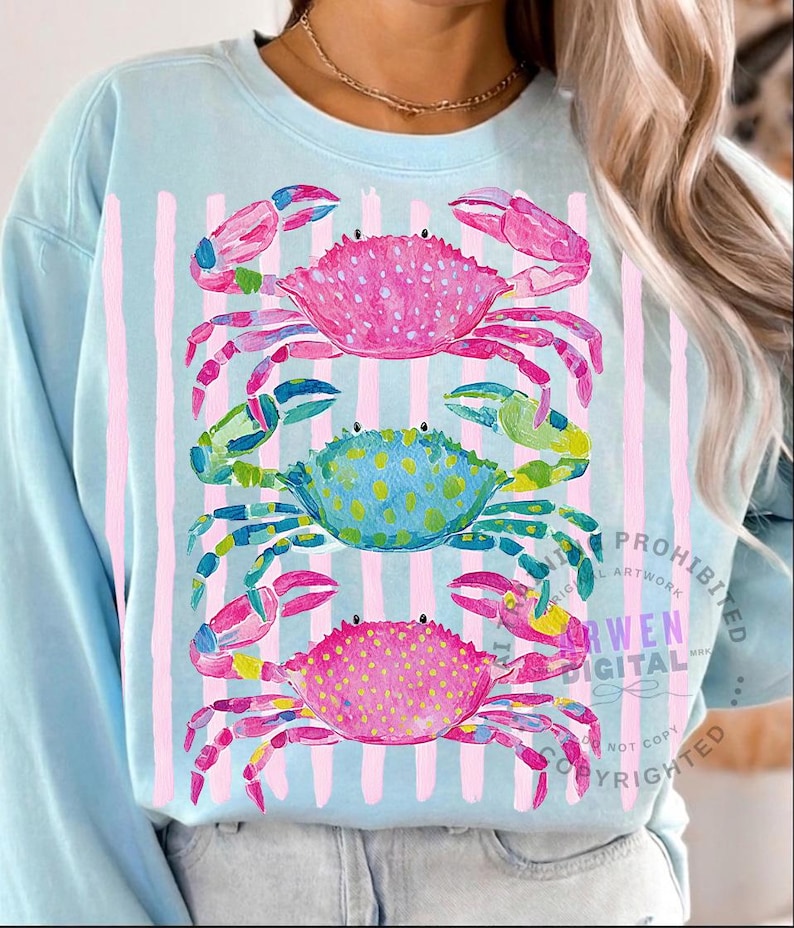 May include: Light blue sweatshirt featuring a watercolor design of three colorful crabs stacked vertically. The crabs are pink, blue, and pink again, with pink and green accents. Vertical pink stripes are behind the crabs.