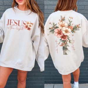 May include: A beige sweatshirt with a floral cross design on the back and the text "JESUS is the light walk by faith not by sight" and the text "JESUS is the light" on the front.