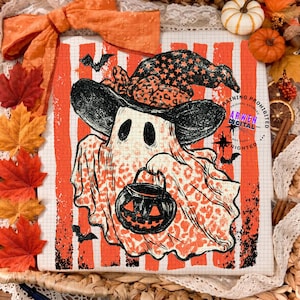 May include: A white t-shirt featuring a graphic of a ghost wearing a witch hat and holding a jack-o'-lantern bucket. The ghost has a leopard print pattern, set against a background of vertical orange stripes and a grid pattern with small bats.