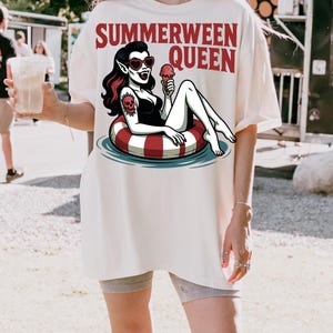 May include: Cream-colored t-shirt with the text "SUMMERWEEN QUEEN" in red. The graphic features a vampire in a black swimsuit, heart-shaped sunglasses, and a skull tattoo, lounging on a red and white striped inner tube, holding an ice cream cone.