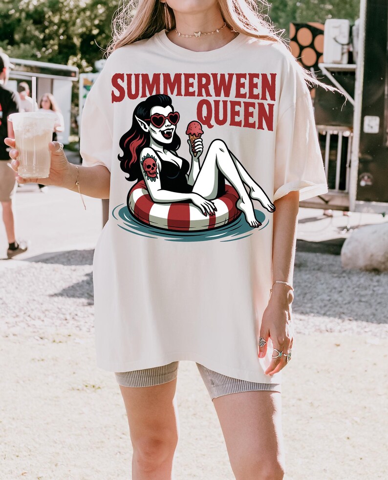 May include: Cream-colored t-shirt with the text "SUMMERWEEN QUEEN" in red. The graphic features a vampire in a black swimsuit, heart-shaped sunglasses, and a skull tattoo, lounging on a red and white striped inner tube, holding an ice cream cone.