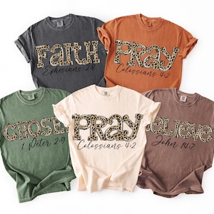 May include: Five coloured t-shirts with inspirational words in leopard print. The shirts are in shades of grey, orange, green, cream, and brown. The words "Faith", "Pray", "Chosen", and "Believe" are printed on the shirts, along with Bible verses.