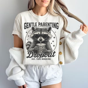 Puede incluir: Camiseta color crema con un mapache con gafas de sol y una bebida. La camiseta tiene el texto "GENTLE PARENTING DROPOUT EST. THIS MORNING" en negro, con una fuente de estilo vintage. El gr&aacute;fico del mapache es en blanco y negro.