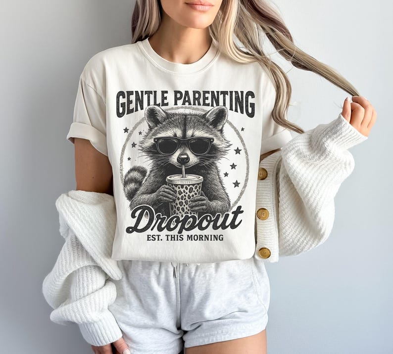 Puede incluir: Camiseta color crema con un mapache con gafas de sol y una bebida. La camiseta tiene el texto "GENTLE PARENTING DROPOUT EST. THIS MORNING" en negro, con una fuente de estilo vintage. El gr&aacute;fico del mapache es en blanco y negro.
