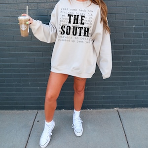May include: A woman wearing a light gray sweatshirt with the text "y'all come back now fixinto hissy fit let THE slower th u go pretty masses each gimme some sugar SOUTH heavens to betsy gussied up jeet yet" printed on the front. She is wearing white high-top sneakers and holding a coffee cup.