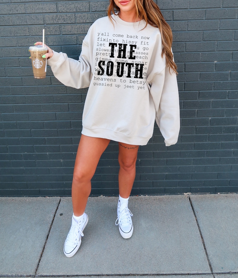 May include: A woman wearing a light gray sweatshirt with the text "y'all come back now fixinto hissy fit let THE slower th u go pretty masses each gimme some sugar SOUTH heavens to betsy gussied up jeet yet" printed on the front. She is wearing white high-top sneakers and holding a coffee cup.