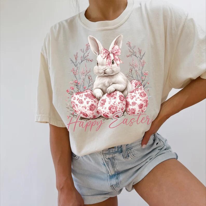 May include: A white t-shirt with a graphic of a white bunny rabbit sitting on pink and white floral patterned Easter eggs. The text "Happy Easter" is printed in pink below the bunny.