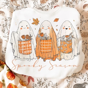 May include: White sweatshirt featuring three cartoon ghosts with smiling faces and orange floral pockets. The ghosts are holding roses and surrounded by falling leaves. The words "Spooky Season" are written in orange script.