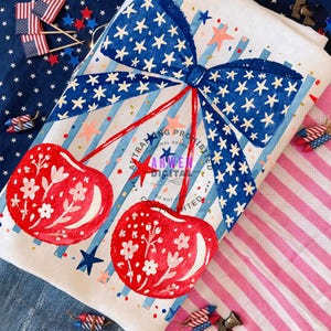 Puede incluir: Un paño de cocina blanco con un diseño patriótico que presenta un lazo azul con estrellas blancas, cerezas rojas y rayas azules y rojas. Las cerezas tienen detalles florales blancos. La toalla está decorada con estrellas y purpurina.