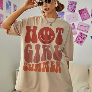 May include: A beige oversized t-shirt with a graphic print of "HOT GIRL SUMMER" in a retro font with a red and orange color gradient. The graphic also includes a smiling face.