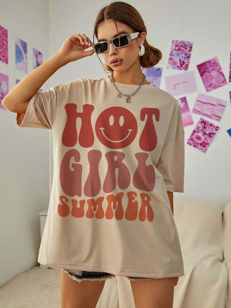 May include: A beige oversized t-shirt with a graphic print of "HOT GIRL SUMMER" in a retro font with a red and orange color gradient. The graphic also includes a smiling face.