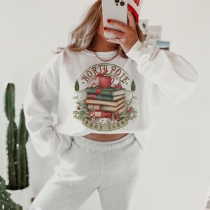 May include: White sweatshirt with a graphic featuring a stack of books, a mug, and the words "North Pole Book Club." The design is surrounded by festive elements. The person is wearing a white and red hat.