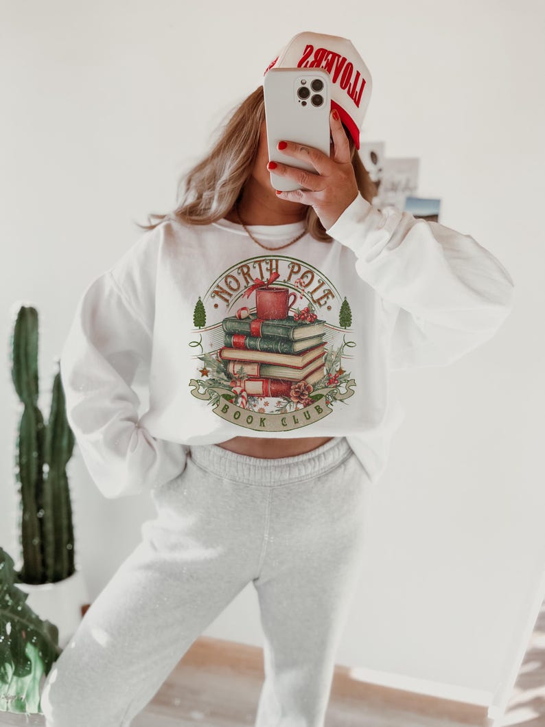 May include: White sweatshirt with a graphic featuring a stack of books, a mug, and the words "North Pole Book Club." The design is surrounded by festive elements. The person is wearing a white and red hat.