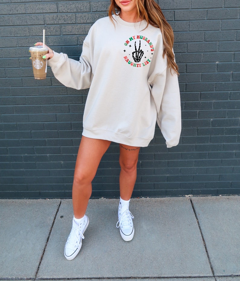 May include: A beige sweatshirt with the text "ON MY HUSBAND'S NAUGHTY LIST" in a circle on the upper left chest. The design includes a skeleton hand making a peace sign. The sweatshirt is paired with white sneakers.