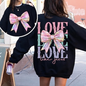 K&ouml;nnte beinhalten: Schwarzer Pullover mit einer glitzernden rosa Schleife und dem Text "LOVE LIKE LOVE like jesus" auf der R&uuml;ckseite.