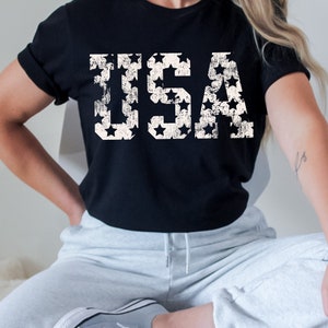 K&ouml;nnte beinhalten: Ein schwarzes T-Shirt mit einem wei&szlig;en, abgenutzten Aufdruck des Wortes "USA", das aus Sternen besteht.