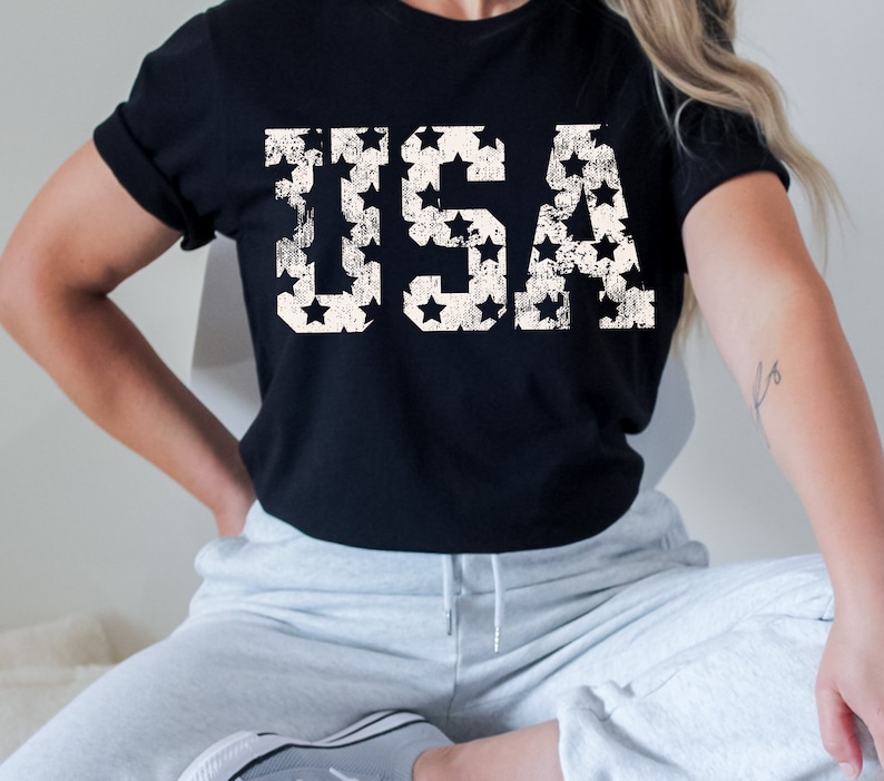 K&ouml;nnte beinhalten: Ein schwarzes T-Shirt mit einem wei&szlig;en, abgenutzten Aufdruck des Wortes "USA", das aus Sternen besteht.