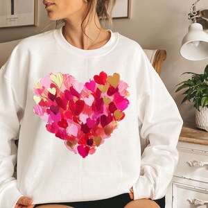 May include: White sweatshirt with a large pink, red, and gold heart design made of many smaller hearts. The heart design is centered on the front of the sweatshirt.