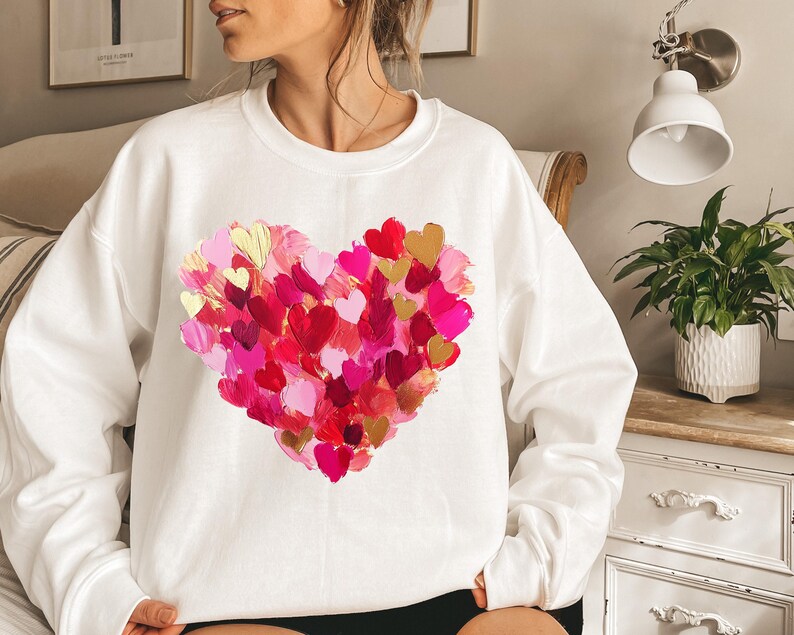 May include: White sweatshirt with a large pink, red, and gold heart design made of many smaller hearts. The heart design is centered on the front of the sweatshirt.