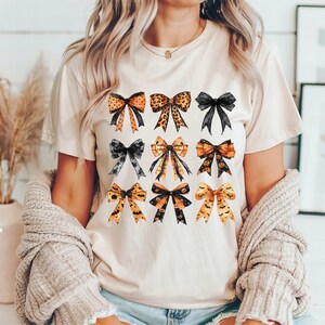 May include: A beige t-shirt featuring a collection of nine watercolor bows in various styles and colors. The bows include leopard print, black, and orange with Halloween-themed designs like bats and pumpkins. The shirt is worn by a person.