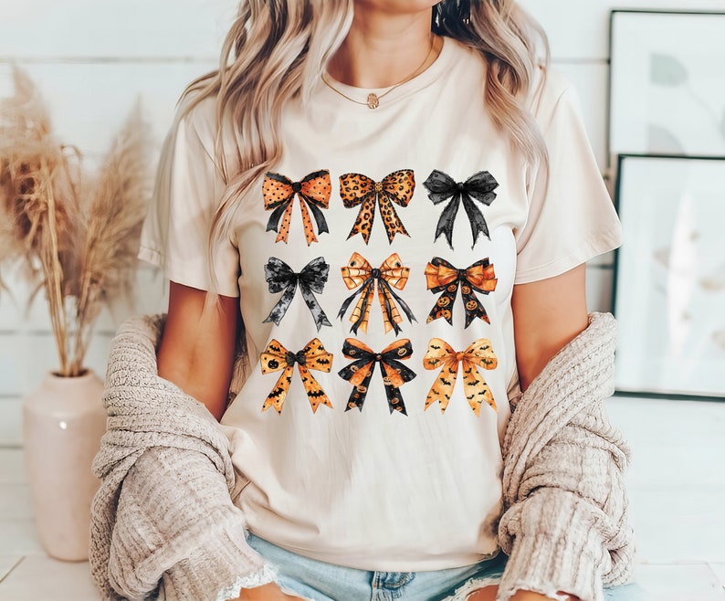 May include: A beige t-shirt featuring a collection of nine watercolor bows in various styles and colors. The bows include leopard print, black, and orange with Halloween-themed designs like bats and pumpkins. The shirt is worn by a person.