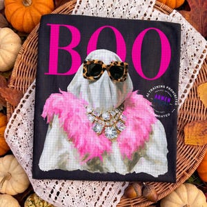May include: A black t-shirt featuring the word BOO in large hot pink letters. The graphic shows a white ghost wearing leopard print sunglasses, a bright pink feather boa, and a large, multi-faceted clear crystal statement necklace.