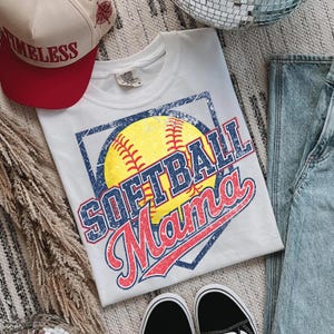 May include: White t-shirt with a distressed graphic of a yellow softball, the words "SOFTBALL Mama" in blue and red, and a blue square border. A red and tan baseball cap with the word "TIMELESS" is also visible.