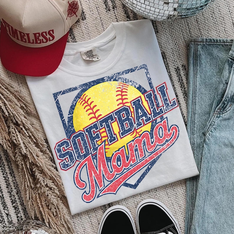 May include: White t-shirt with a distressed graphic of a yellow softball, the words "SOFTBALL Mama" in blue and red, and a blue square border. A red and tan baseball cap with the word "TIMELESS" is also visible.