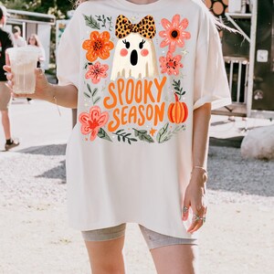 K&ouml;nnte beinhalten: Cremefarbenes &uuml;bergro&szlig;es T-Shirt mit einem Halloween-Design. Es zeigt einen Geist mit Leopardenmuster-Schleife, umgeben von orangefarbenen und rosa Blumen, und den Worten "Spooky Season". Ein K&uuml;rbis und Sterne vervollst&auml;ndigen das Design.