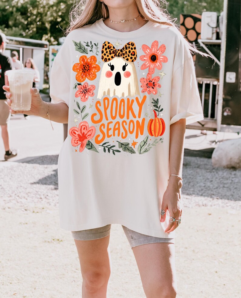 K&ouml;nnte beinhalten: Cremefarbenes &uuml;bergro&szlig;es T-Shirt mit einem Halloween-Design. Es zeigt einen Geist mit Leopardenmuster-Schleife, umgeben von orangefarbenen und rosa Blumen, und den Worten "Spooky Season". Ein K&uuml;rbis und Sterne vervollst&auml;ndigen das Design.