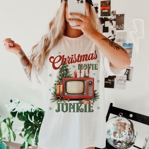 May include: White t-shirt with a Christmas-themed graphic. The design features the words "Christmas Movie Junkie" in red and green, along with a vintage television, Christmas tree, and gifts. The person in the image is wearing a black baseball cap.