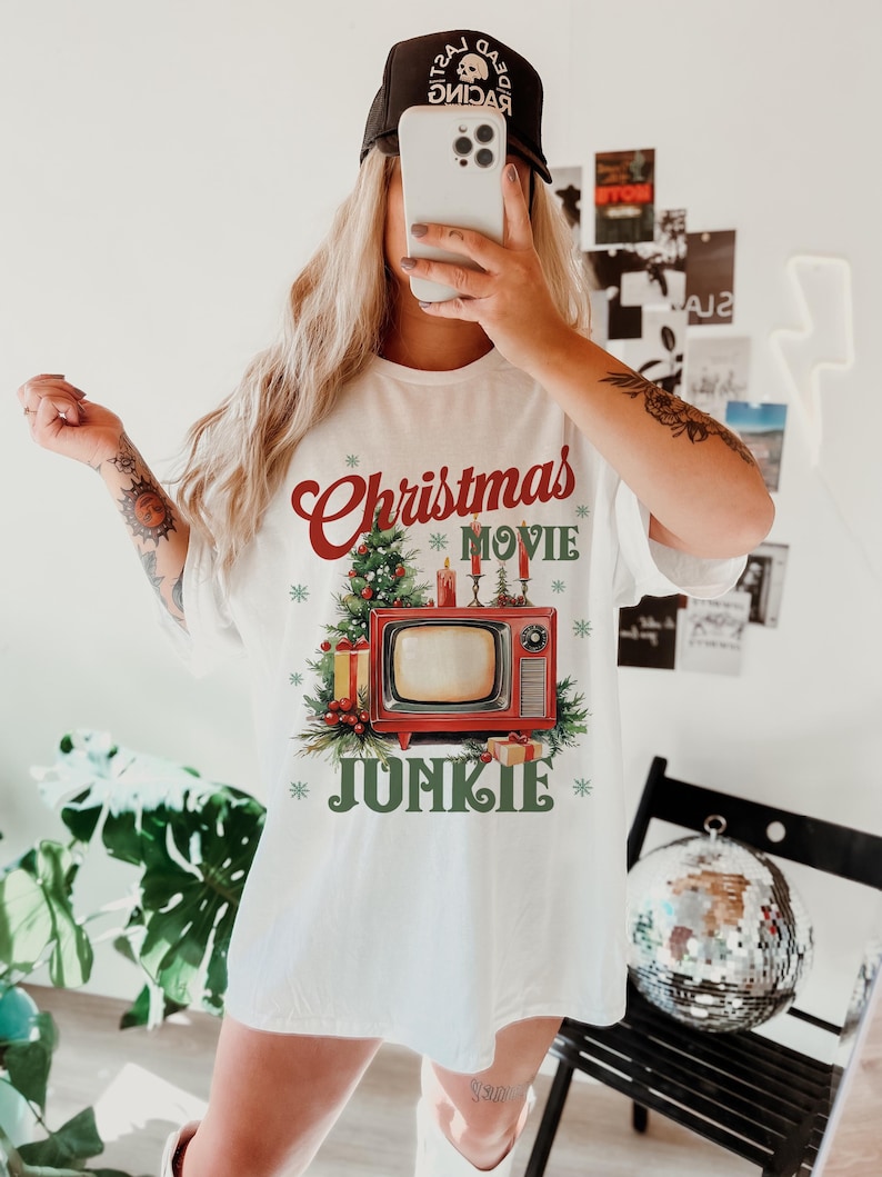 May include: White t-shirt with a Christmas-themed graphic. The design features the words "Christmas Movie Junkie" in red and green, along with a vintage television, Christmas tree, and gifts. The person in the image is wearing a black baseball cap.