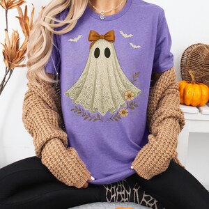 May include: A purple t-shirt featuring a ghost design with a brown bow, two black eyes, and a lace-like body. The ghost is surrounded by floral and bat graphics. The shirt is part of a Halloween-themed collection.