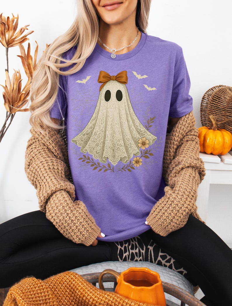 May include: A purple t-shirt featuring a ghost design with a brown bow, two black eyes, and a lace-like body. The ghost is surrounded by floral and bat graphics. The shirt is part of a Halloween-themed collection.
