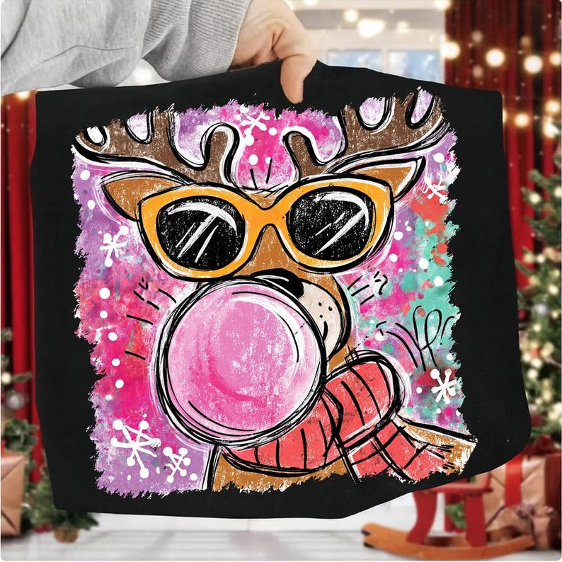 Op de afbeelding: Zwart T-shirt met een cartoon rendier met een zonnebril die een kauwgomballenblaas blaast. Het rendier heeft bruine geweien en een rood-zwarte sjaal. De achtergrond heeft roze, paarse en turquoise accenten met sneeuwvlokken.