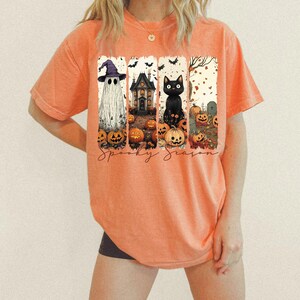 May include: Orange t-shirt with a Halloween-themed graphic. The graphic features a ghost wearing a witch hat, a haunted house, a black cat, pumpkins, and the words "Spooky Season".