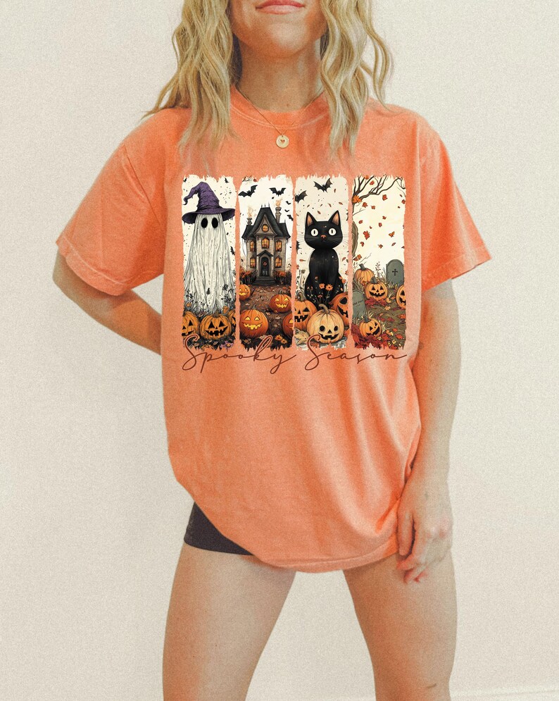 May include: Orange t-shirt with a Halloween-themed graphic. The graphic features a ghost wearing a witch hat, a haunted house, a black cat, pumpkins, and the words "Spooky Season".