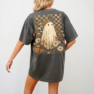 Op de afbeelding: Donkergrijs T-shirt met een spookontwerp en bloemmotieven. De geest heeft zwarte ogen en is omgeven door oranje en witte bloemen. Boven de geest bevindt zich een dambordpatroon.