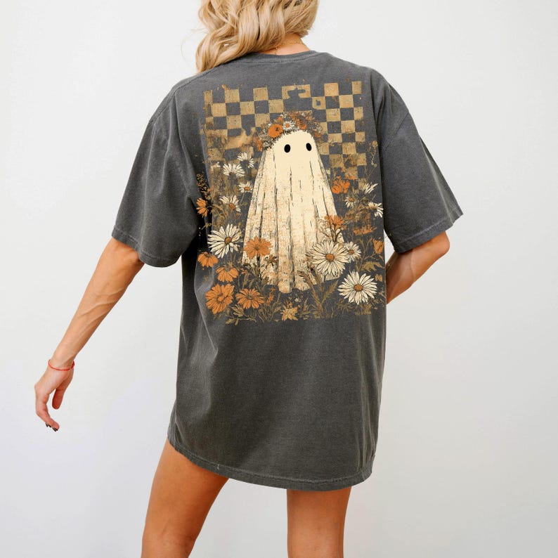 Op de afbeelding: Donkergrijs T-shirt met een spookontwerp en bloemmotieven. De geest heeft zwarte ogen en is omgeven door oranje en witte bloemen. Boven de geest bevindt zich een dambordpatroon.
