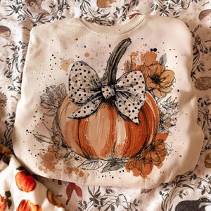 May include: Cream-colored sweatshirt featuring a large orange pumpkin with a black and white polka dot bow. The pumpkin is surrounded by floral accents and brown paint splatters, creating a fall-themed design.