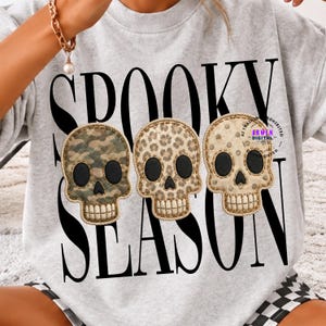 Peut inclure: Un sweat-shirt gris clair à col rond avec le texte noir SPOOKY SEASON et trois motifs de crânes. Les crânes sont disposés horizontalement, chacun avec un motif différent : camouflage vert, imprimé léopard marron et pois beiges.