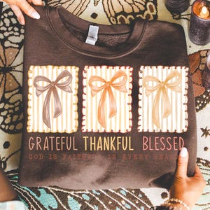 May include: A brown sweatshirt featuring three rectangular designs with watercolor bows in shades of brown, orange, and beige. The text "Grateful Thankful Blessed" is printed below the designs, with the phrase "God is faithful in every season" underneath.