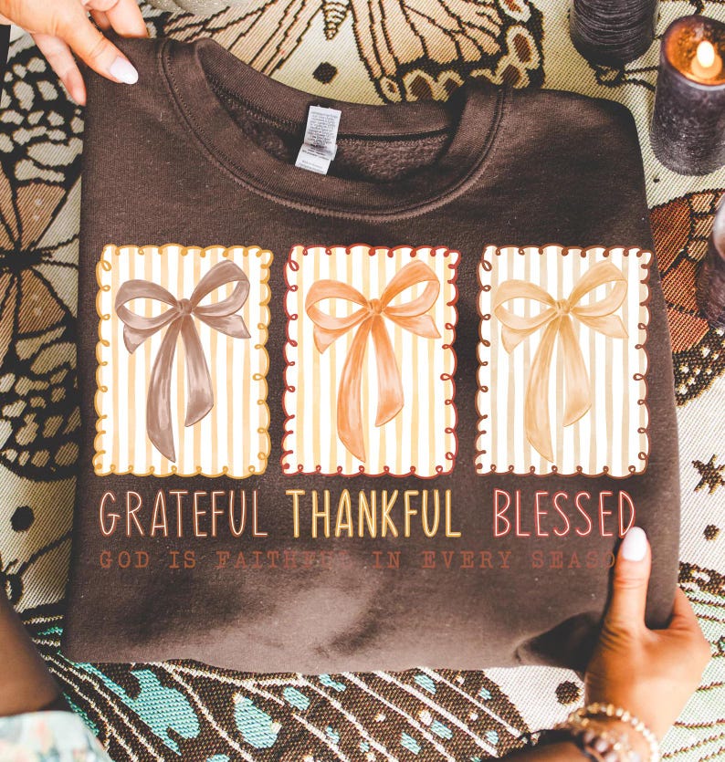 May include: A brown sweatshirt featuring three rectangular designs with watercolor bows in shades of brown, orange, and beige. The text "Grateful Thankful Blessed" is printed below the designs, with the phrase "God is faithful in every season" underneath.