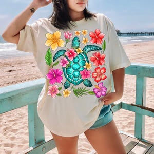 May include: A cream coloured t-shirt with a graphic of a green sea turtle surrounded by pink, red, yellow, and orange hibiscus flowers.